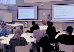 Outi Loikkanen puhumassa verkostotapaamisessa, etualalla näkyy osallistujia istumassa pöytien ääressä.
